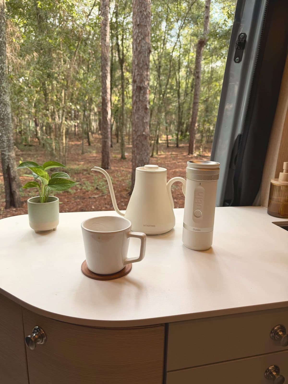 A gooseneck kettle, OutIn grinder and mug on Nellie's galley counter, with pines outside the open door