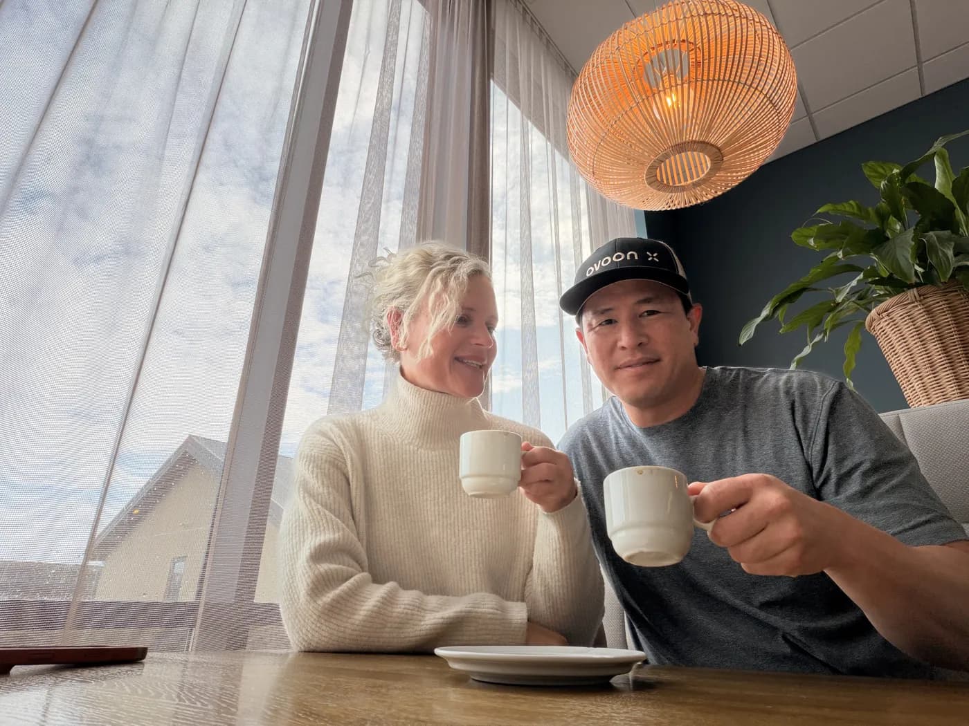 Linsey and Randy raising white coffee cups by a café window under a rattan lamp