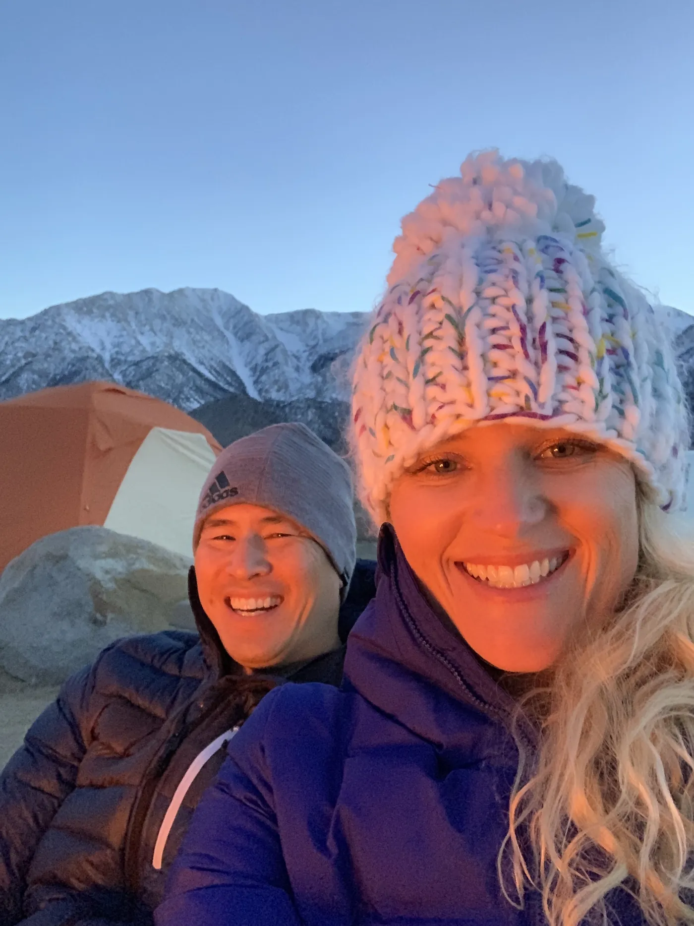 Randy and Linsey smiling in warm firelight at camp, with an orange tent and snowy mountains behind them